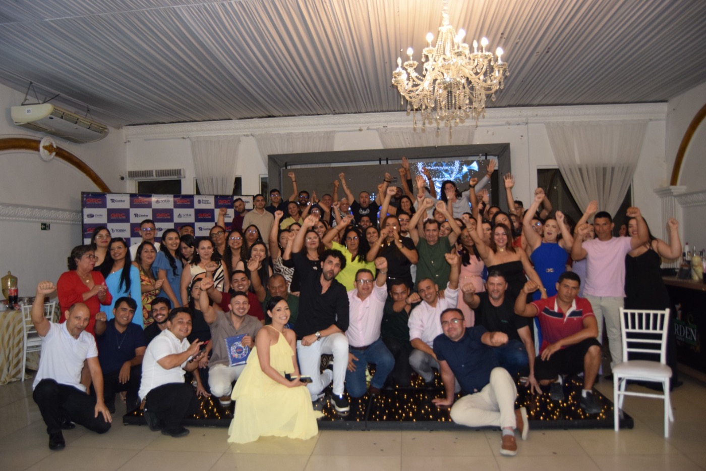 Equipe do Grupo R Center reunida em uma confraternização, comemorando de braços erguidos diante de um painel com os logos GRC, R Center, Torra, Super Fraldas e Vita