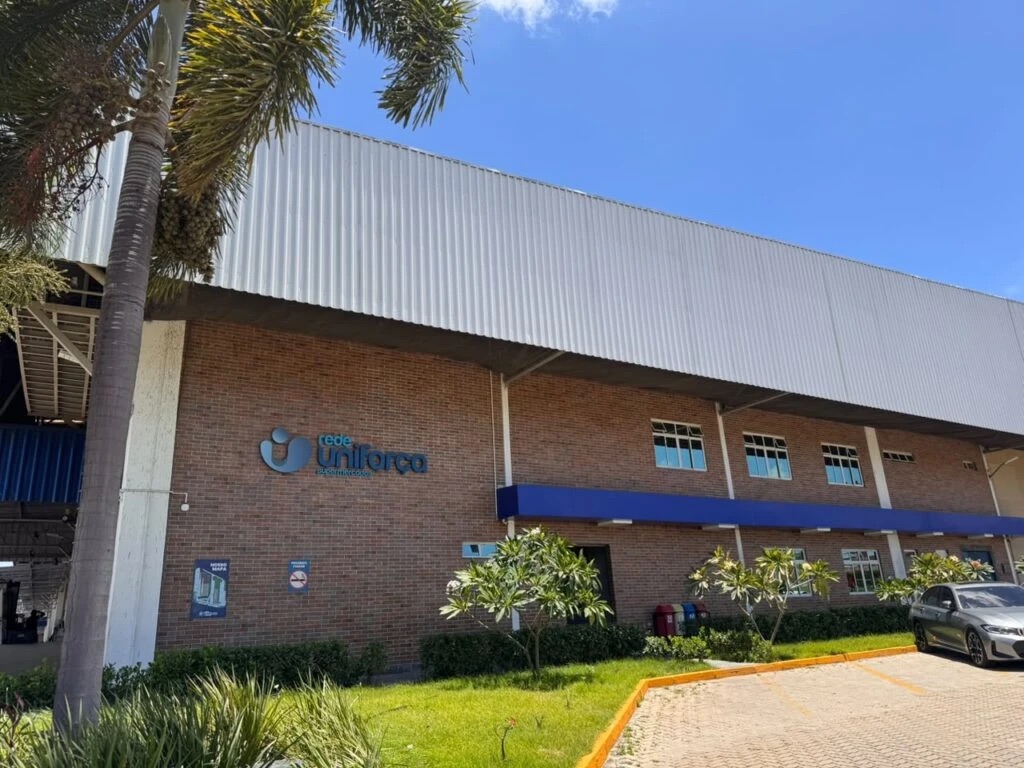 Fachada do centro de distribuição da Rede Uniforça, prédio de tijolinhos com o logo da rede, palmeiras e jardim na frente